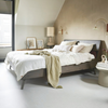 Rustige slaapkamer met zandkleurige accentmuur, strak grijs bedframe, frisse witte vloer en subtiele decoratie zoals ingelijste prints, lampen en een beige katoenen plaid.