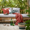 Tuinlounge met bamboe tuinset, gestyled met terracotta kussen, gebloemd sierkussen en glazen waterflessen op een jute buitenkleed en omringd door veel groene planten.