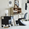 Scandinavische werkkamer met licht houten bureau, open vakken voor opberging, zwarte S-vormige designstoel en minimalistisch zwart ladenkastje op wieltjes, op een wit gelakte houten vloer.