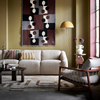 Lichte woonkamer met beige bouclé bank, retro houten fauteuil en abstract wandkleed in aardetinten, gecombineerd met gele designlamp en geometrisch vloerkleed.